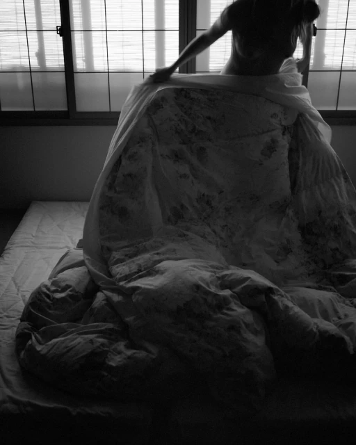Monochrome photograph of a person holding up a patterned duvet cover in front of a window. Light filtering through window.