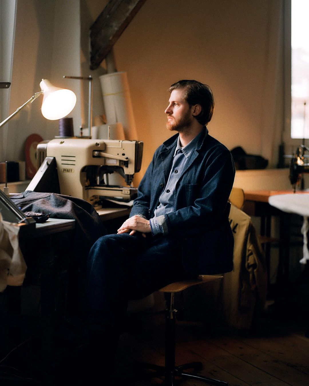 Man in denim jacket seated near sewing machine in workshop. Tailoring scene with natural light, fabric, and classic sewing tools.