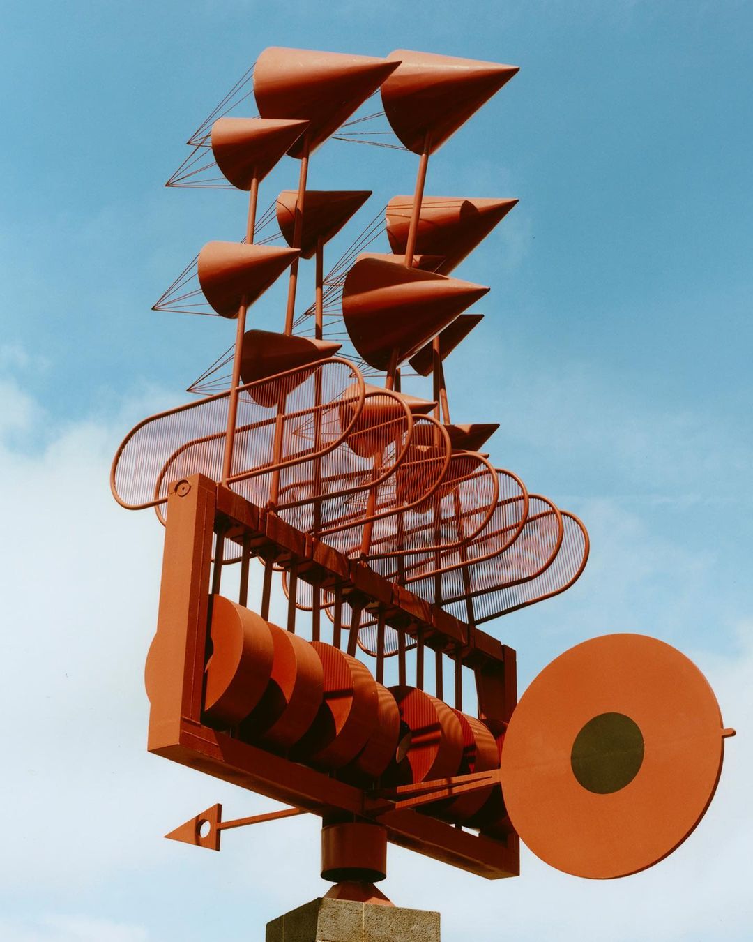 Red metal sculpture featuring geometric shapes and a wind vane against a bright blue sky. Abstract art on a stone pedestal.