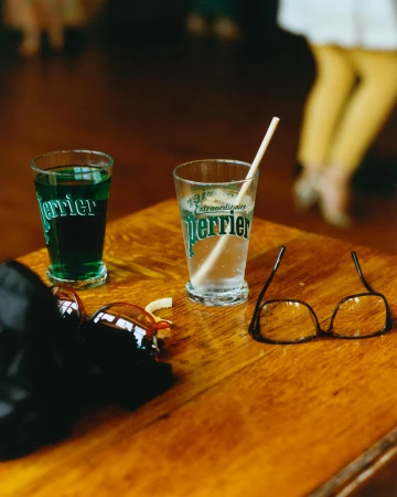 Two Perrier glasses, one with a straw, sit on a wooden table next to glasses, a hat, and the legs of a person are in the background.
