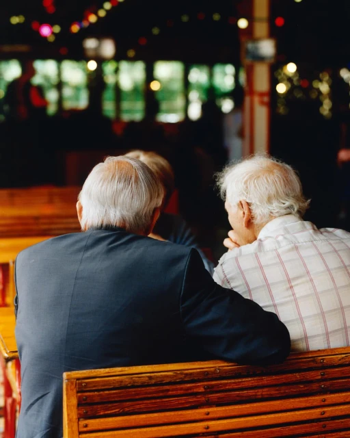 Two elderly men sitting on a wooden bench at an outdoor cafe, viewed from behind, with a dark interior and colorful lights in background.