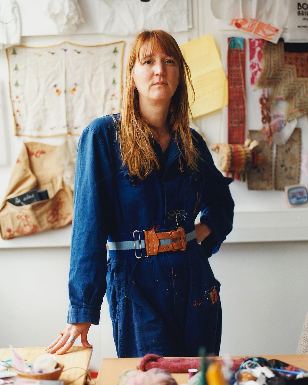 Red-haired woman in a blue jumpsuit stands in a studio with crafting supplies, sewing tools, and vintage textiles.