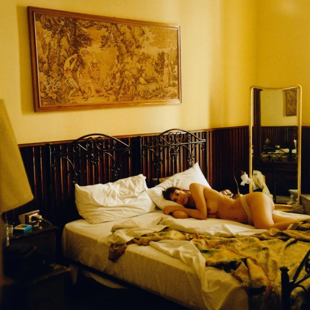 Nude woman lying on bed in an indoor room, decorated with an ornate painting, mirror, and wrought iron bedframe.  
