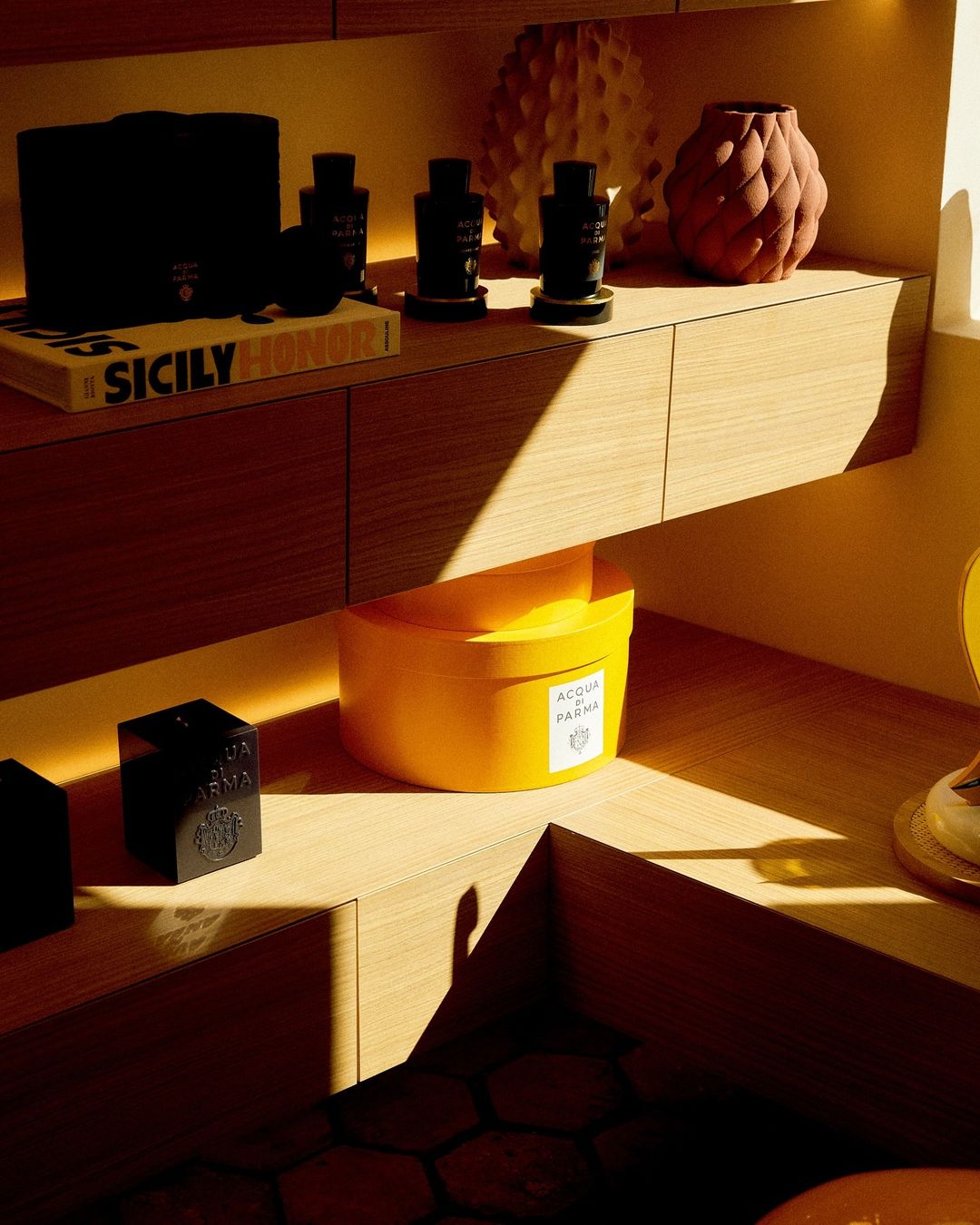 Close-up shot of perfume bottles and decorative objects on wooden shelves, featuring 'Sicily' book, warm lighting, and textured backdrop.