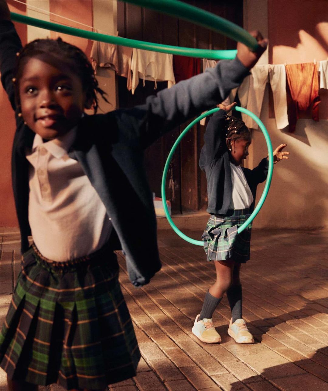 Two young Black girls in school uniforms play with green hula hoops outdoors on a brick surface with clothes hanging to dry in the background.