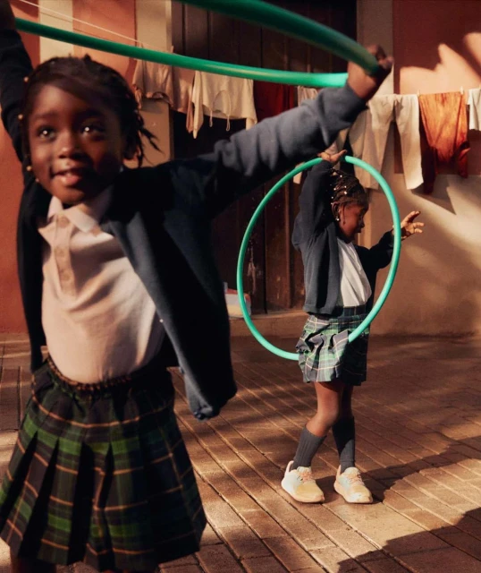 Two young Black girls in school uniforms play with green hula hoops outdoors on a brick surface with clothes hanging to dry in the background.
