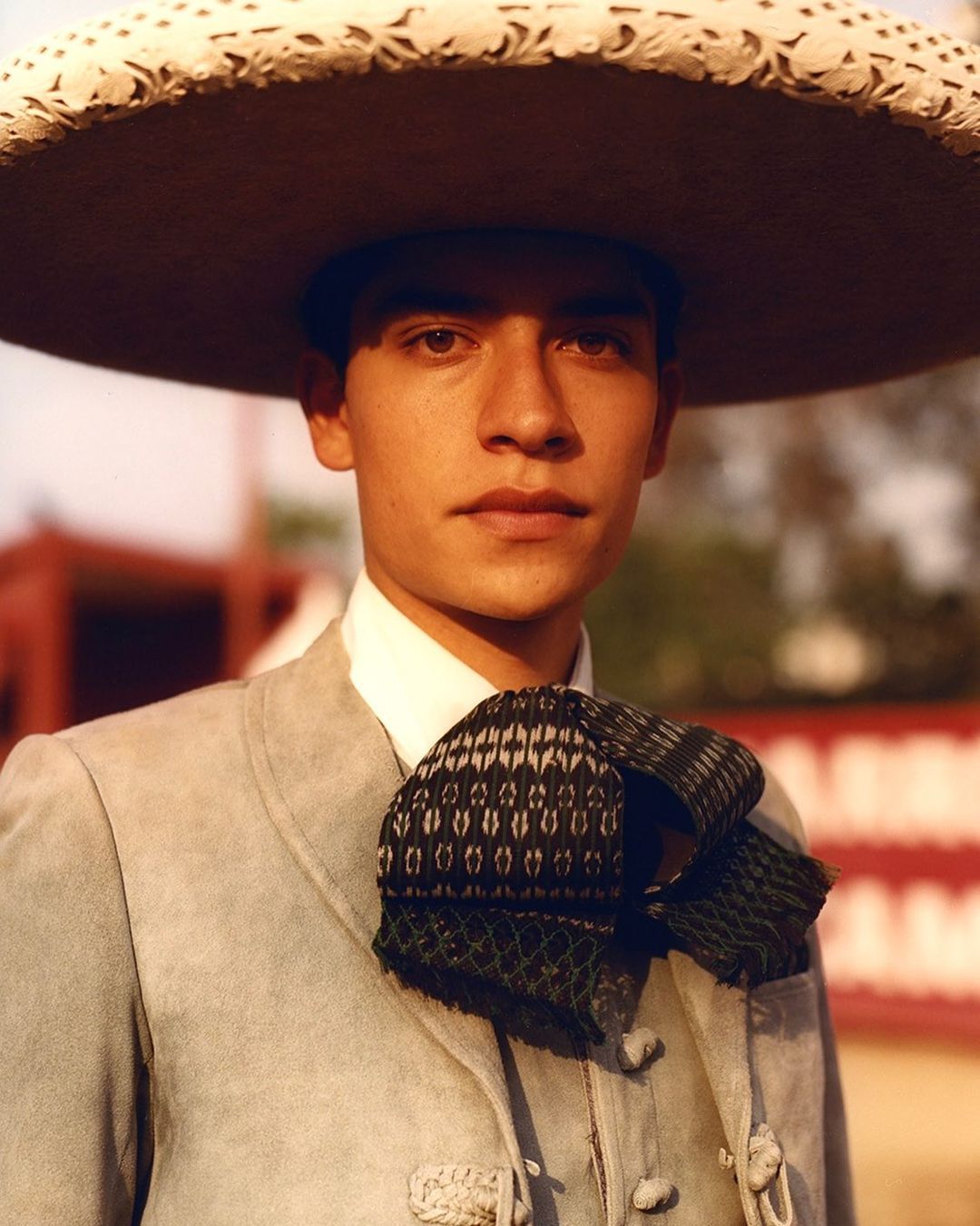 A young man in traditional Mexican charro attire poses in a close-up shot, wearing a large sombrero and patterned ascot, warm lighting.