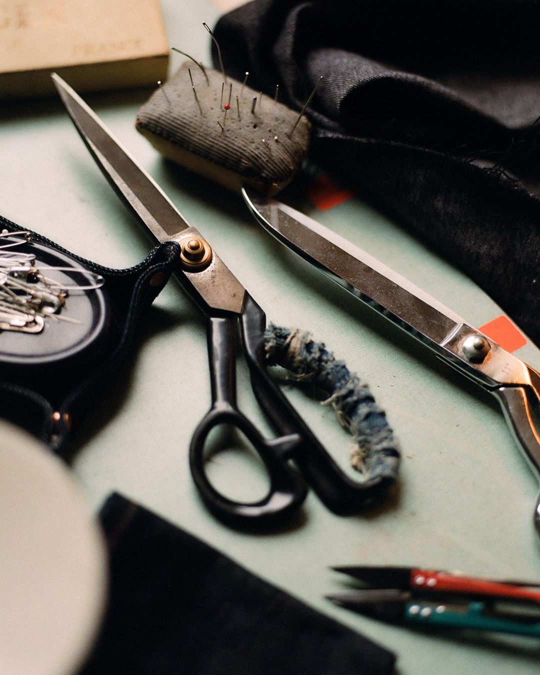Close-up still life of sewing tools including scissors, pins in a pin cushion, and black cloth on a light green surface.