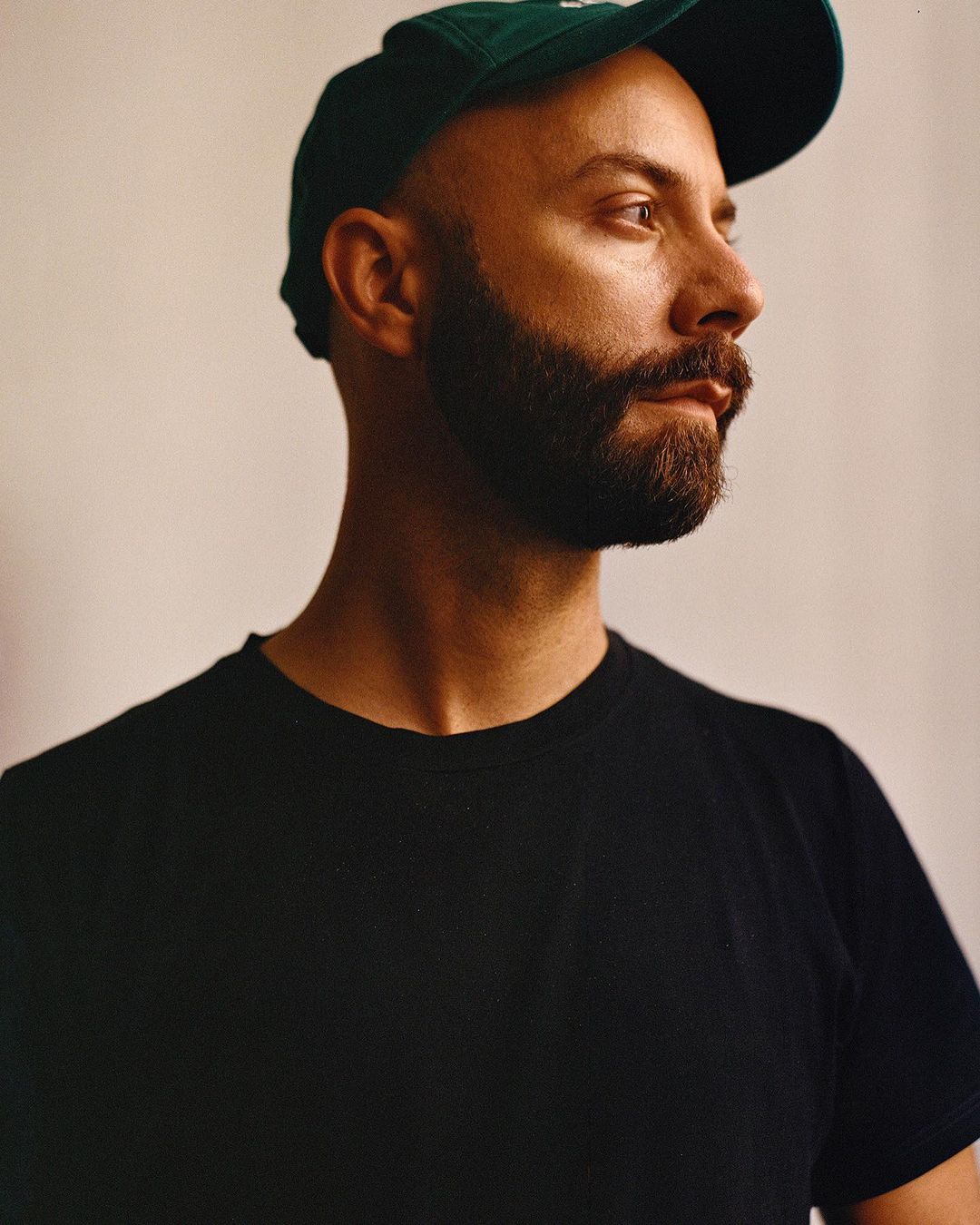 Headshot portrait of a man wearing a green baseball cap and a black t-shirt against a neutral studio backdrop, looking away from camera.