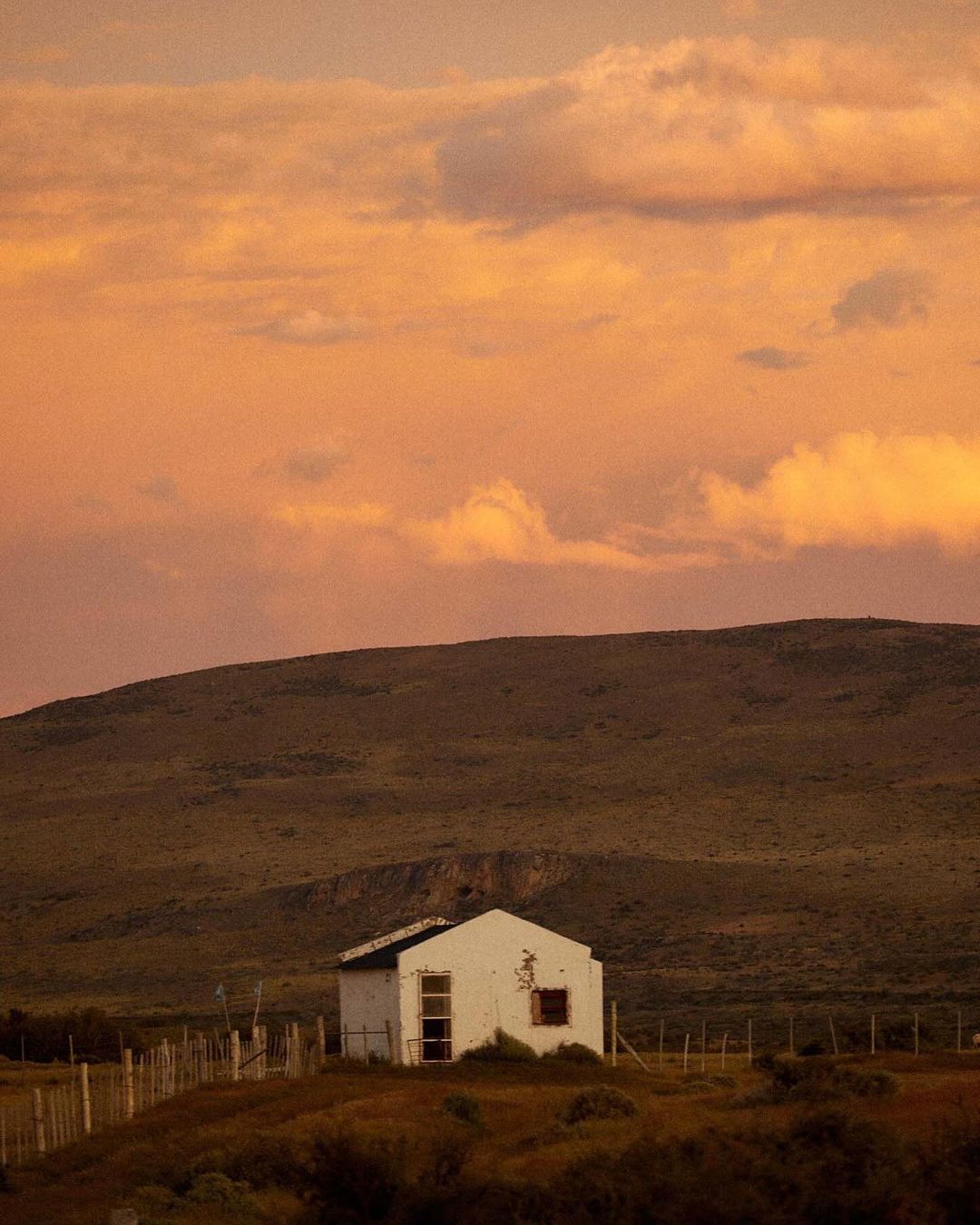 Dilapidated white building in a rural landscape with rolling hills and a fiery orange sunset sky with clouds, evoking a serene atmosphere.