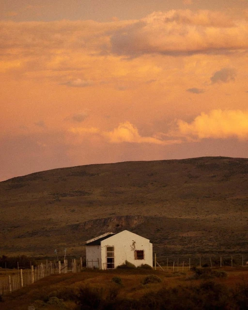 Dilapidated white building in a rural landscape with rolling hills and a fiery orange sunset sky with clouds, evoking a serene atmosphere.