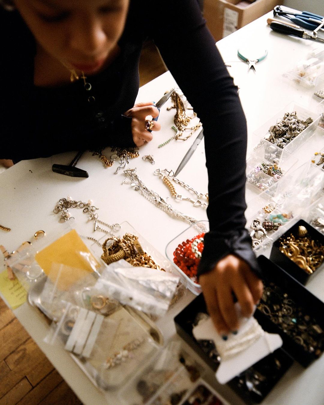 Woman crafts jewelry at white table with bead containers, tools, and chain necklaces in an indoor setting, close up, high angle.