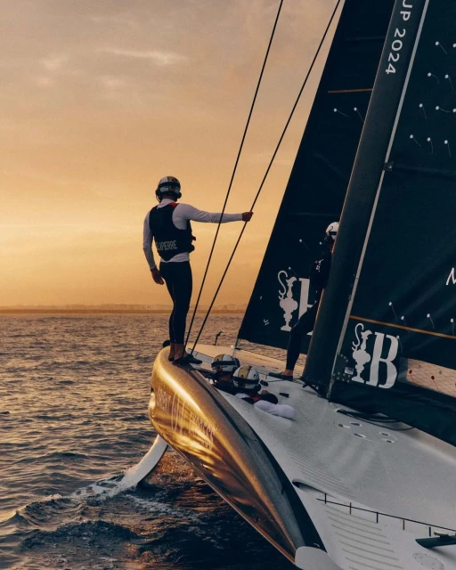 Sailboat racing at sunset with two sailors on deck wearing helmets and life vests, sailing through choppy water with a golden sky.
