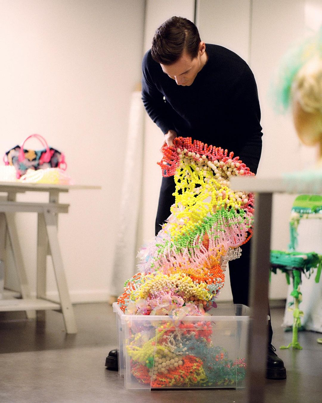 Adult male organizing a multicolored beaded sculpture into a clear plastic bin, displayed in a bright art studio, fashion, and design.