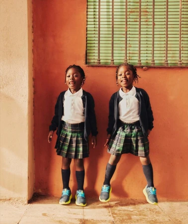 Two young Black girls stand outdoors against a bright orange wall in their school uniforms, consisting of pleated skirts, white shirts, and cardigans.