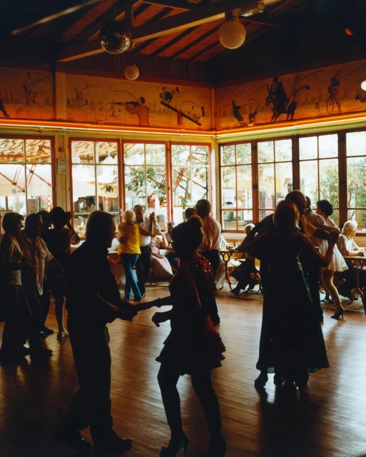 Silhouetted figures ballroom dancing in a softly lit, high-ceilinged room with large windows and a disco ball hanging from the ceiling.