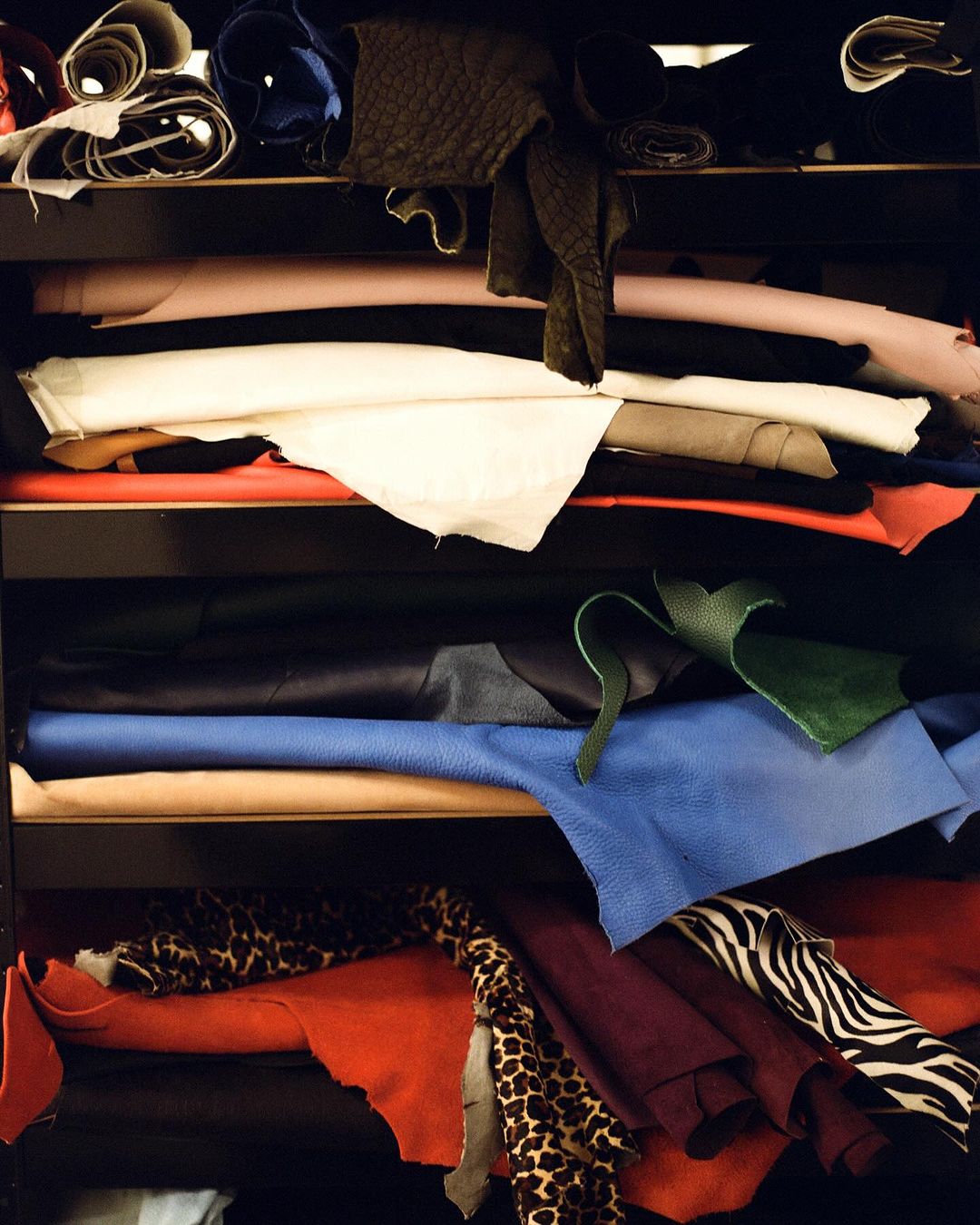 Shelves filled with colorful piles of leather and patterned fabric in rolls and draped, with textures and colors creating a dynamic composition.