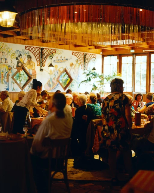 Indoor restaurant scene with many people seated at tables, served by staff, under decorative lighting with mural wall.