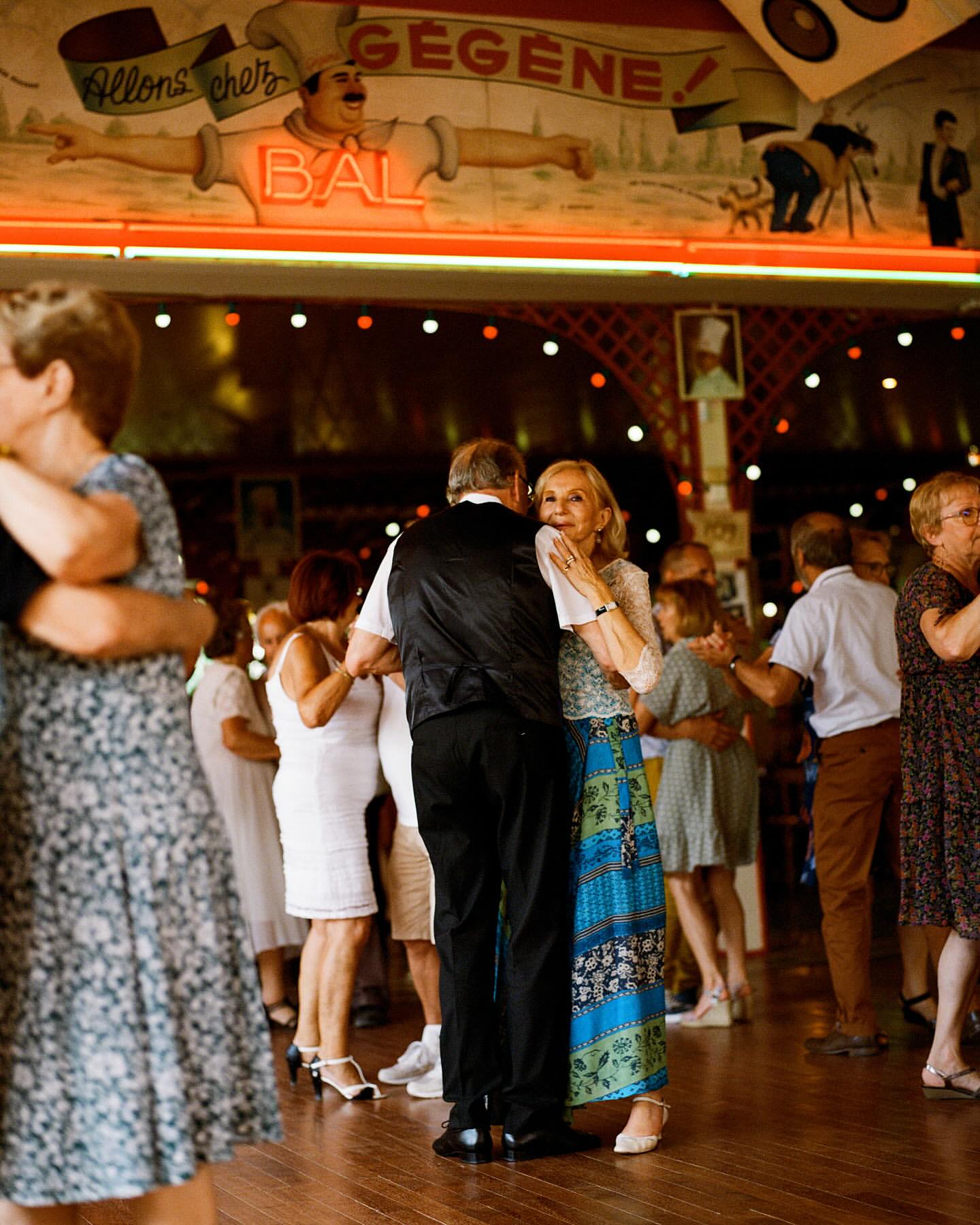Several couples of adult people are dancing together at a ball room with a mural of a man in the background. The wooden floor is shiny.