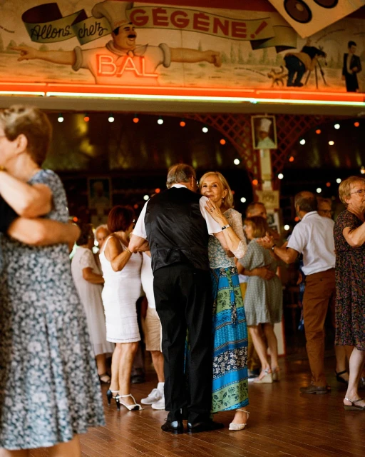 Several couples of adult people are dancing together at a ball room with a mural of a man in the background. The wooden floor is shiny.