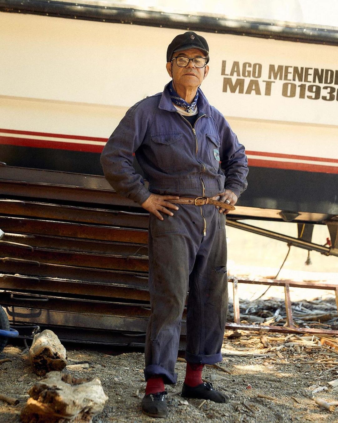 Older man wearing coveralls, cap, and glasses stands in front of a boat and stacked metal sheets in an outdoor setting.