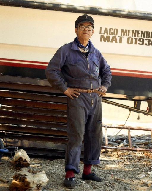 Older man wearing coveralls, cap, and glasses stands in front of a boat and stacked metal sheets in an outdoor setting.