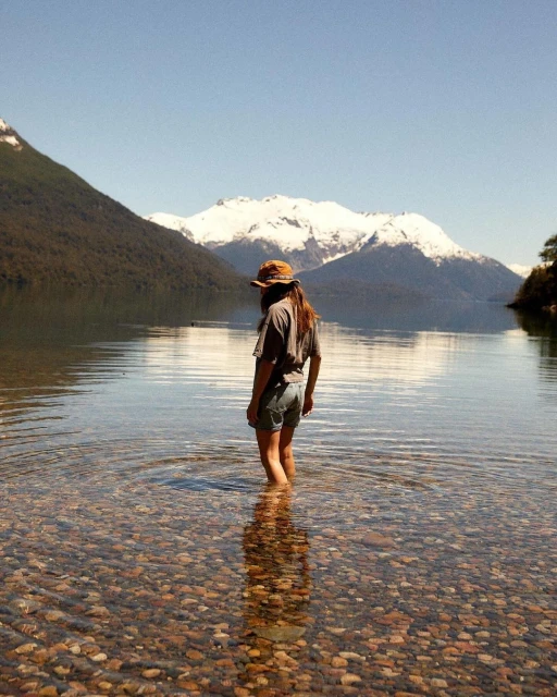 A woman stands in a clear, shallow lake with mountains in the background, wearing shorts, a t-shirt, and a hat on a sunny day.