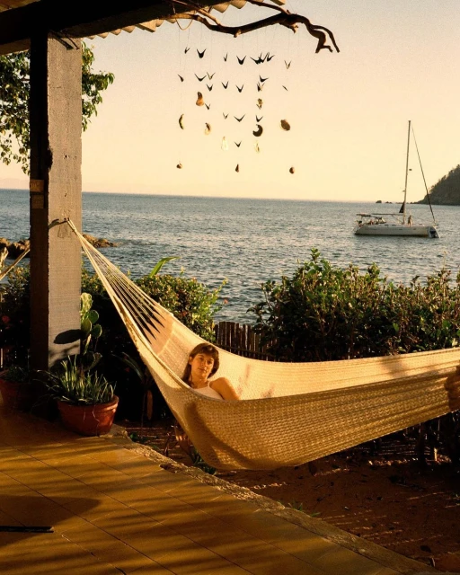 Woman relaxing in a hammock on a porch with ocean view, sailboat in the distance, under decorative mobile with plant life nearby.