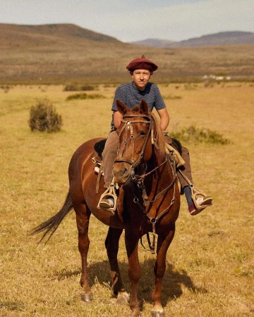 A young person with beret rides a chestnut horse through grassy plains in the countryside on a sunny day, showcasing an equine scene.