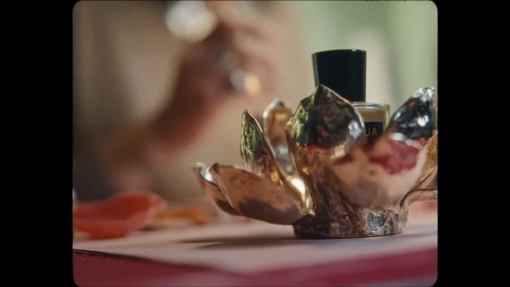 Close-up shot of a perfume bottle in a gold lotus-shaped holder, with a blurred person in the background, against a softly blurred background.