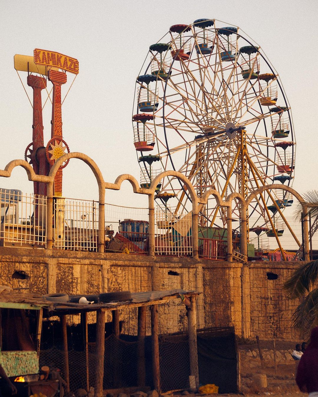 A Ferris wheel and Kamikaze ride are set against a pale sky, appearing in an amusement park with weathered architecture.