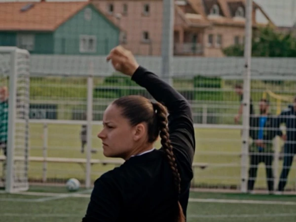 Adult female soccer player prepares for a game on an outdoor field. Green grass and a blurred stadium in the background.