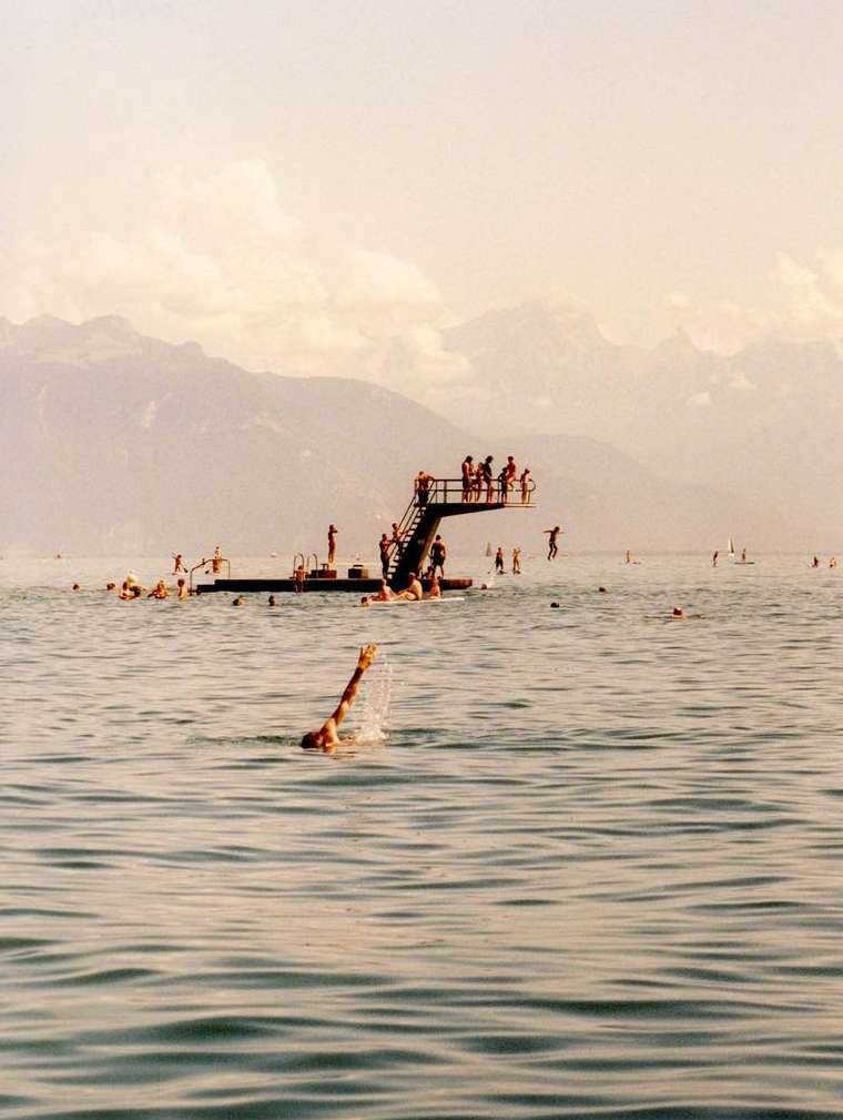 People swim and jump into a blue lake near a diving platform, with mountains in the distance under a sunny sky, creating a summertime scene.