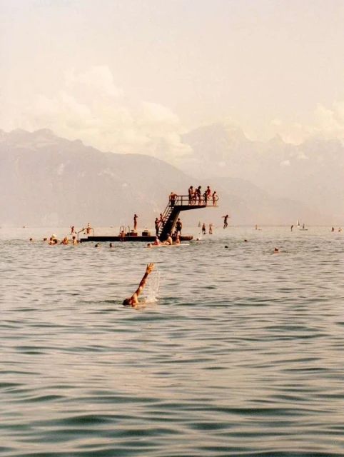 People swim and jump into a blue lake near a diving platform, with mountains in the distance under a sunny sky, creating a summertime scene.