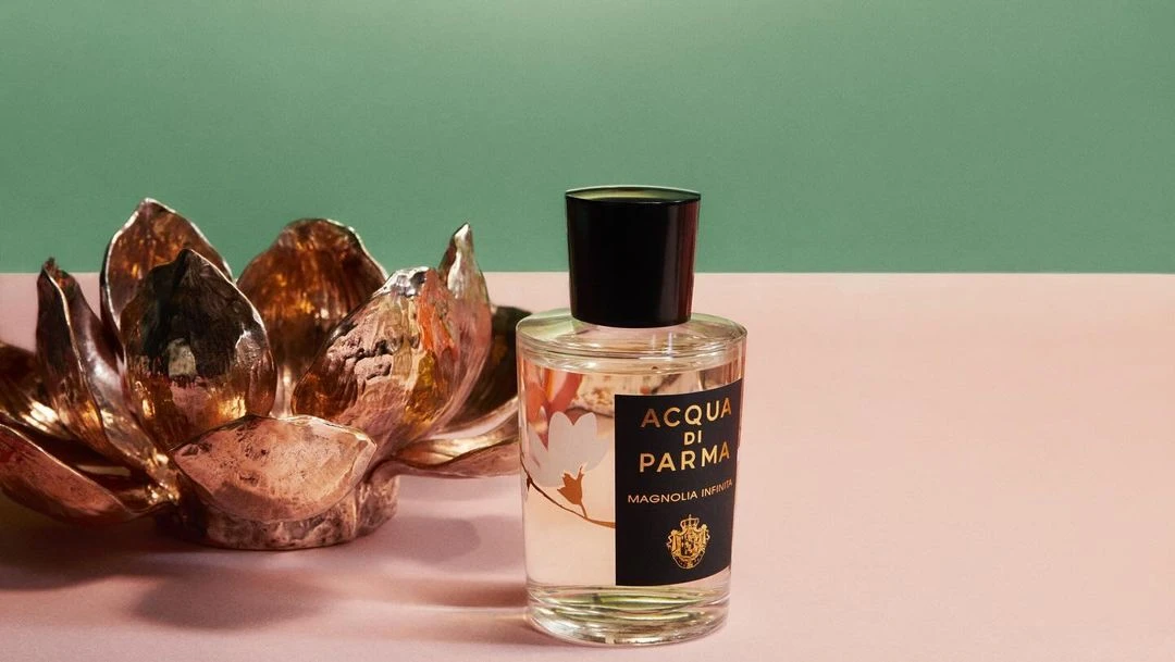Acqua di Parma perfume bottle with a black cap and gold label, resting on a peach surface next to a bronze decorative lotus flower.