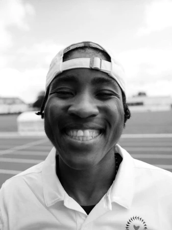 Close-up monochrome portrait of a smiling Black athlete wearing a white baseball cap and polo shirt outdoors.