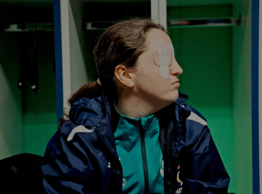 Adult woman with athletic wear sits in a locker room with a bandage over her eye in a medium shot with green and blue lockers.