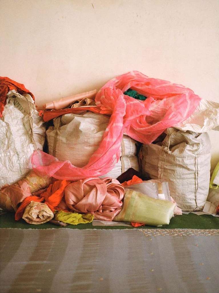 Assorted textiles, including a pink scarf, are piled atop bulk bags indoors against a pale wall, creating a colourful, textured composition.
