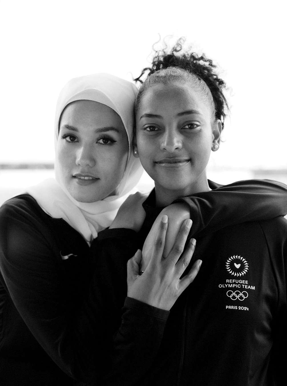 Two smiling women, one wearing a hijab, pose closely in a black and white portrait; Olympic Refugee Team logo visible on clothing.