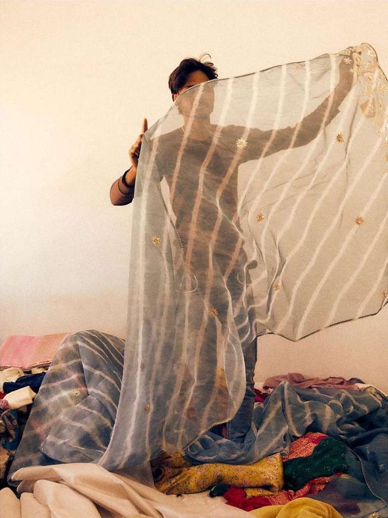 A person stands partially hidden behind a semi-transparent striped fabric, with a pile of colorful textiles beneath it indoors.