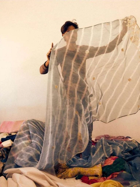 A person stands partially hidden behind a semi-transparent striped fabric, with a pile of colorful textiles beneath it indoors.