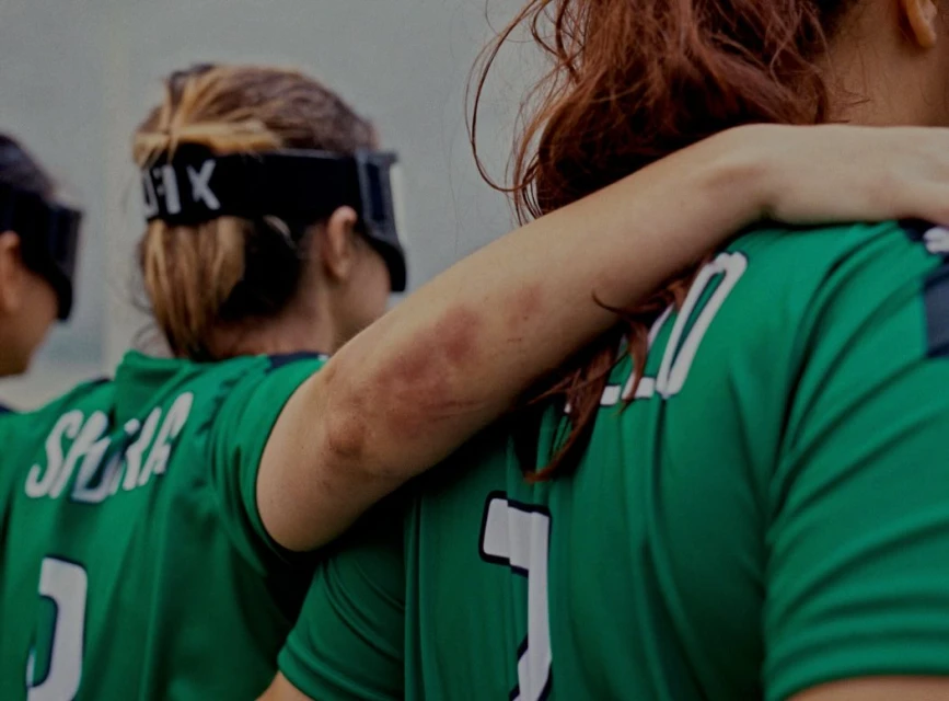 Close shot of female athletes in matching green jerseys with numbers, arms around each other, wearing protective eyewear.