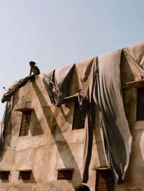 Woman sits on a weathered building with textiles draped over the edge, contrasting with the clear sky above in a sunny, outdoor setting.