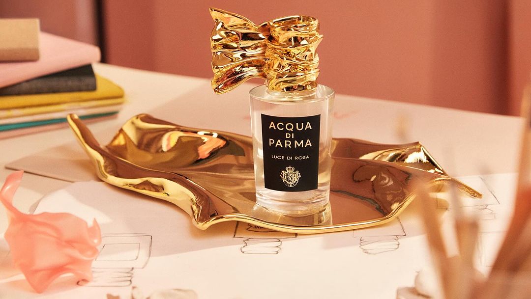 Acqua di Parma Luce di Rosa perfume bottle with a gold decorative bow on a gold leaf-shaped tray, surrounded by desk accessories.