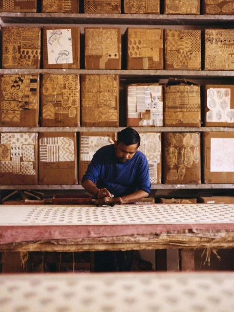Artisan hand-printing textile on long table with patterned wooden blocks on shelves in background. Craftsman working in art studio.