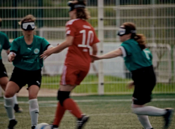 Female soccer players in action on a green field wearing eye covers, one in red jersey and others in green, demonstrating athletic skills.