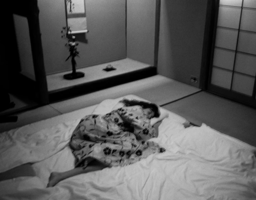 Monochrome shot of a woman asleep on a bed in a room with traditional Japanese decor, featuring tatami mats and a decorative display.