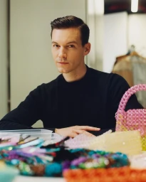 Man with dark hair wearing a black sweater seated behind a table filled with colourful beaded handbags and accessories.