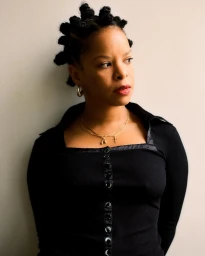 Portrait of a Black woman with Bantu knots wearing a black button-down top and gold jewelry against a pale wall.