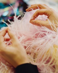 Close-up of hands delicately holding and arranging pale pink feathers in a soft-focus workshop setting.
